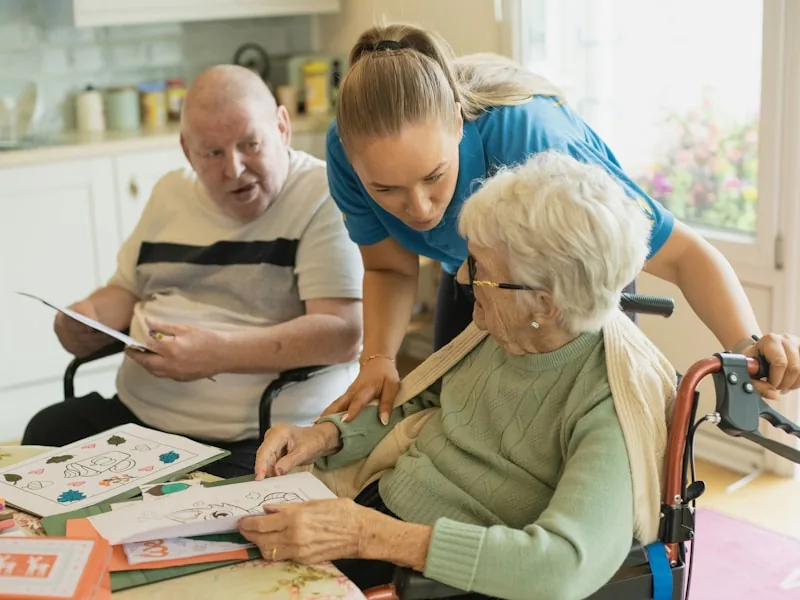 A carer in a blue tunic leans over to help an older woman in a wheelchair with a card-making activity, while an older man reads at the same table