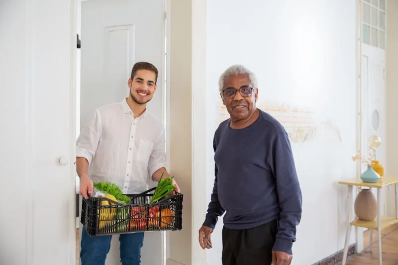 A young carer holding a crate of fresh vegetables at the door of an older man's home, with the man standing in the hallway