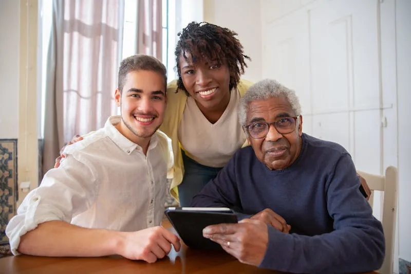 An older man holding a tablet at a table, with a young man and a young woman smiling beside him