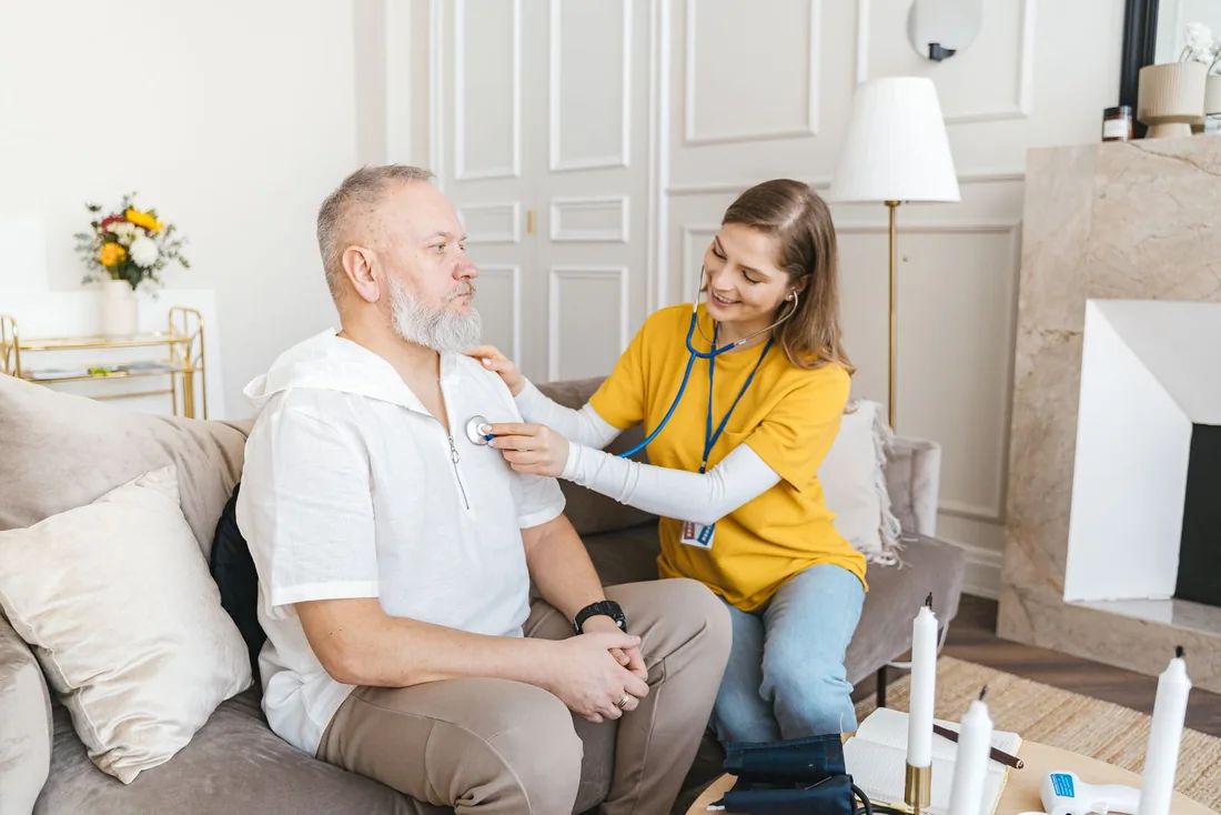 A homecareOS carer checks an older man's health with a stethoscope during a home visit, recording the observation on the app as she goes